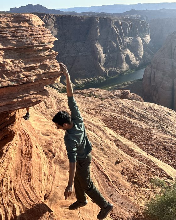 Teja hanging one-handed from a sandstone overhang above a river canyon