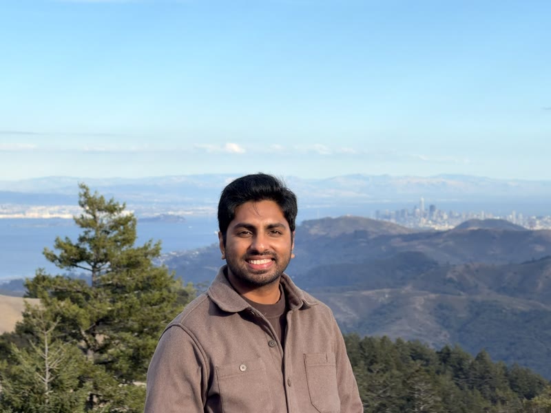 Teja smiling outdoors, with hills, water and a city skyline behind him