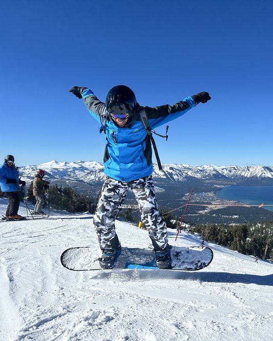 Teja on a snowboard with arms out, above a lake and snowy mountains